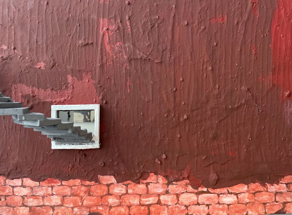 Bild einer roten Backsteinmauer mit einem kleinen Fenster. Durch das Fenster führt eine freischwebende Treppe.